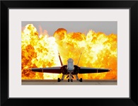 An F/A 18 Hornet sits on the flight line as a wall of fire detonates behind it