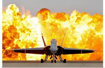 An F/A 18 Hornet sits on the flight line as a wall of fire detonates behind it