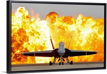 An F/A 18 Hornet sits on the flight line as a wall of fire detonates behind it