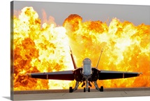 An F/A 18 Hornet sits on the flight line as a wall of fire detonates behind it