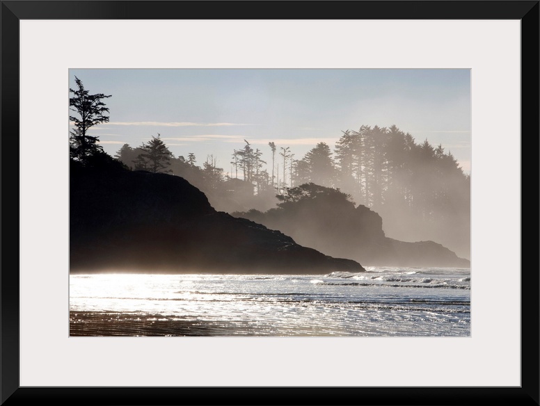 Mist rises of the sea against the silhouettes of rocks and trees in this shore line photograph taken in the morning.