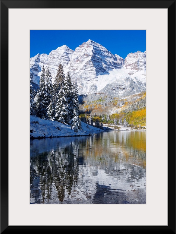 Snow covered mountains are pictured against a clear blue background that reflects down in the body of water that is in the...