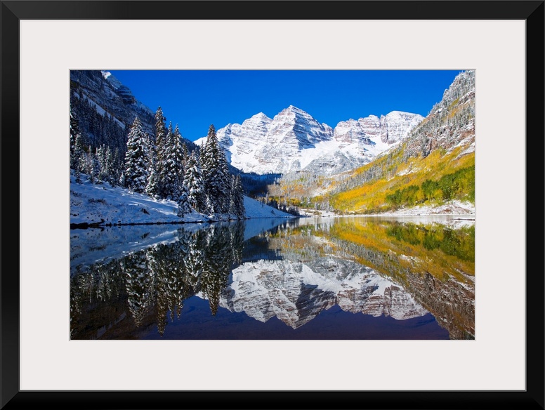 A snow covered mountain range with tall trees in front reflects beautifully into a body of water.