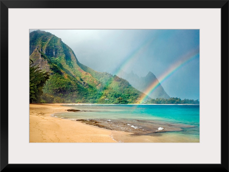 A landscape photograph with double rainbows on a tropical beach with mountains in the background.