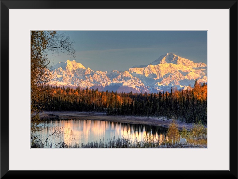 Serene landscape at dawn of two snow-covered mountains near a pine forest at the edge of a small pond in Alaska.