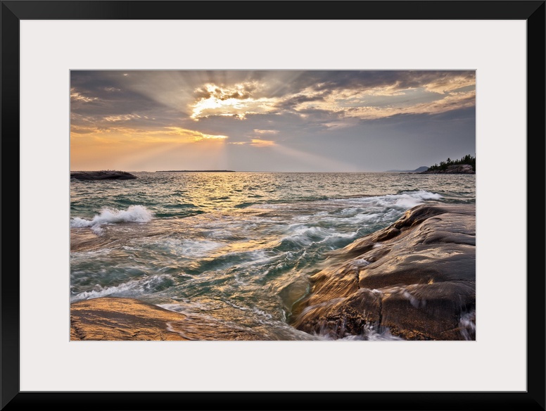Burst of sun rays peeking out behind a cloud as waves lap on to a rocky shore.