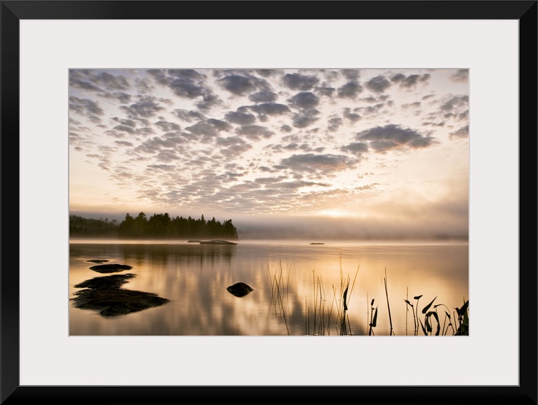 Horizontal photograph on a big wall hanging of the sun set reflecting in a large body of water, a distant tree line in the...