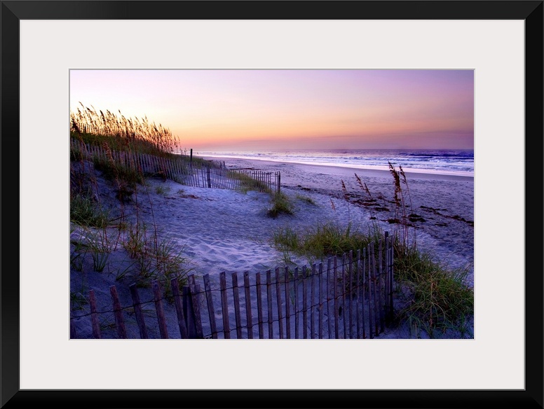 A landscape photograph of a dune covered in sea grass and fences fills the foreground of this beach at sunset.