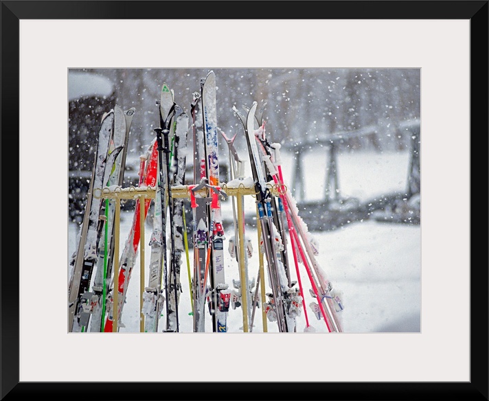 In this photograph multiple skis are stored on an outside rack as large white snowflakes fall all around. Blurred backgrou...