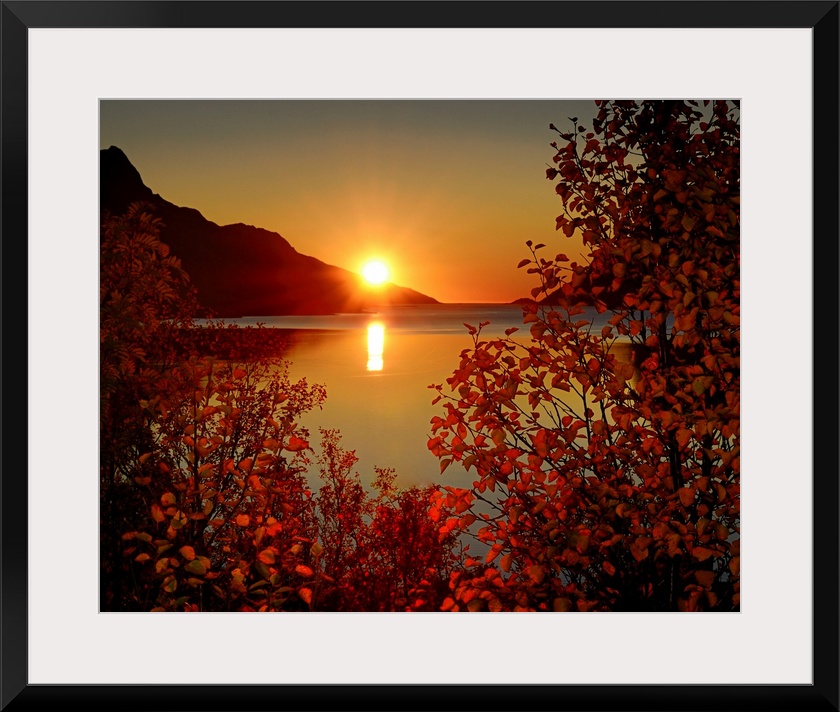 Big photograph emphasizes the sun as it begins to set over a mountain and then reflect over a calm body of water in Norway...