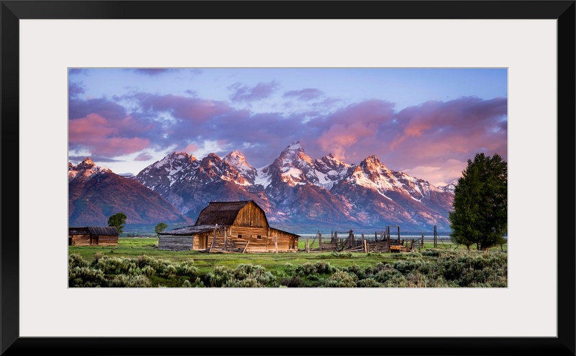 Large, horizontal photograph of an open field with an old, wooden barn and a small building next to that.  Large, snow cap...
