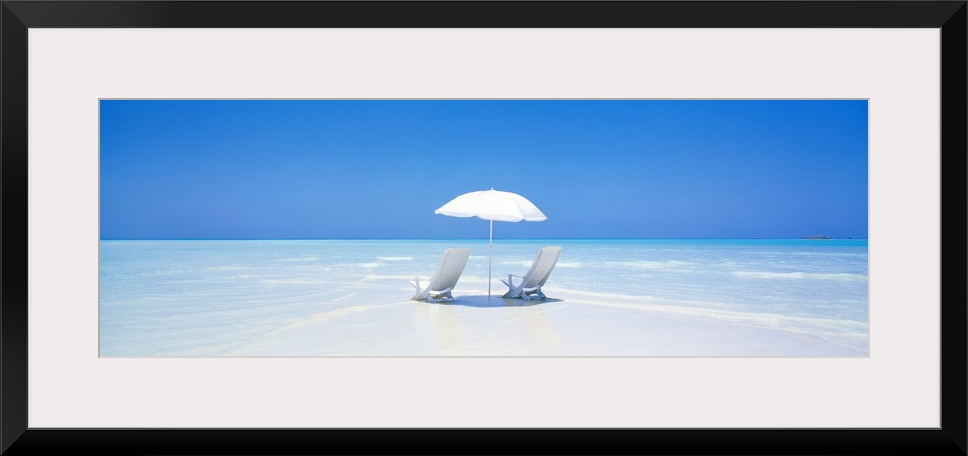 A panoramic photograph displaying two chairs sitting underneath an umbrella on a sandy beach in Maldives.  The clear water...