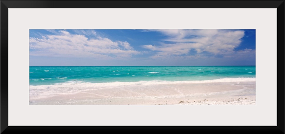 Panoramic photograph of calm ocean with surf and sand in the foreground and cloudy sky above.