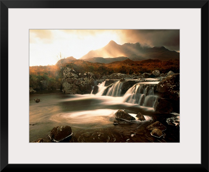 Time lapsed photograph of water flowing through a rocky river while the sun glows behind clouds in the distanced.