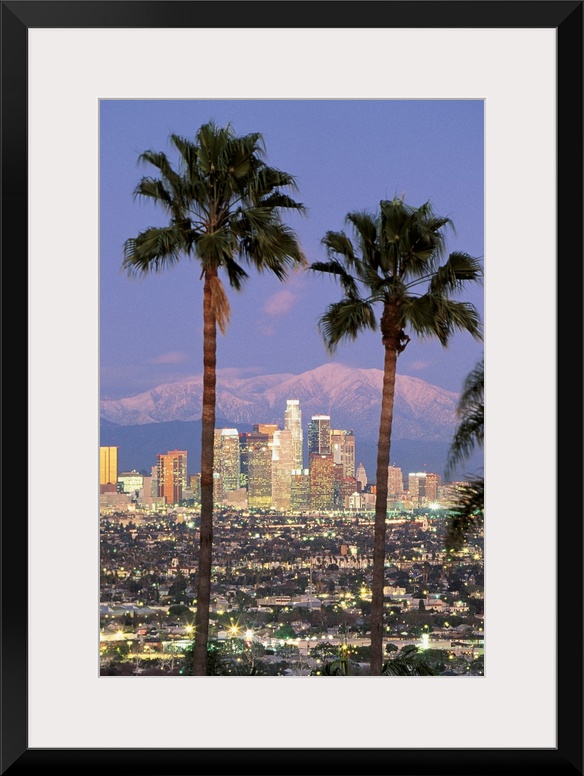 A vertical photograph of skyscrapers in the Los Angelesos skyline framed by two palm trees and mountains in the background.