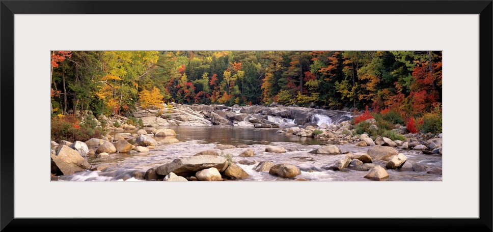 A relaxed panoramic landscape of large boulders in a New England river lined with trees with autumn colors.