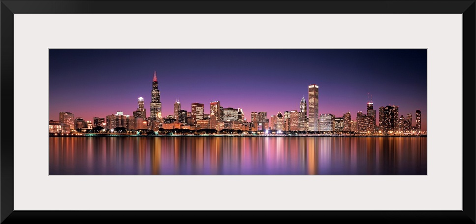 Panoramic photograph of the Chicago, Illinois skyline filled with skyscrapers reflecting onto Lake Michigan at night.