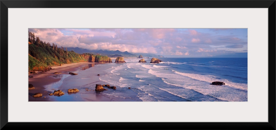 Panoramic photograph of waves slowly crashing against the rocks and sand of Cannon Beach in Oregon.  The mountains in the ...