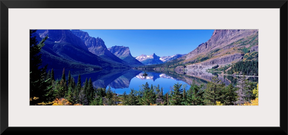 A photographic print of rugged mountains with snow in the distance reflected in a crystal clear lake with foliage in the f...