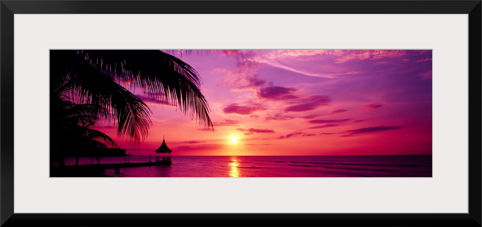 Panoramic photograph of a colorful sunset on the beach in Montego Bay, Jamaica.  The palm trees and dock in the foreground...