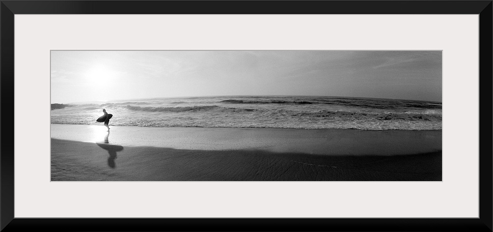 Panoramic photograph of a surfer walking along a sandy beach in San Diego, California.  The tide of the ocean gently crash...