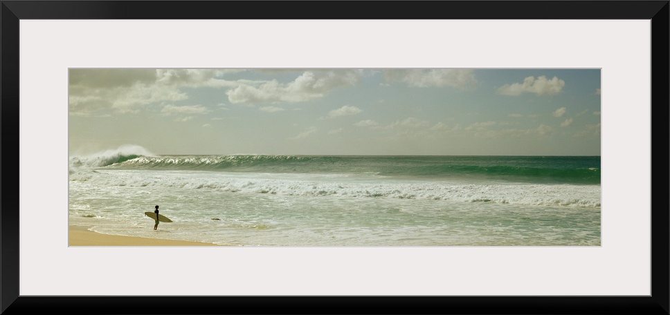 Panoramic image of a surfer standing where the ocean meets the beach shore looking at a big wave crashing.