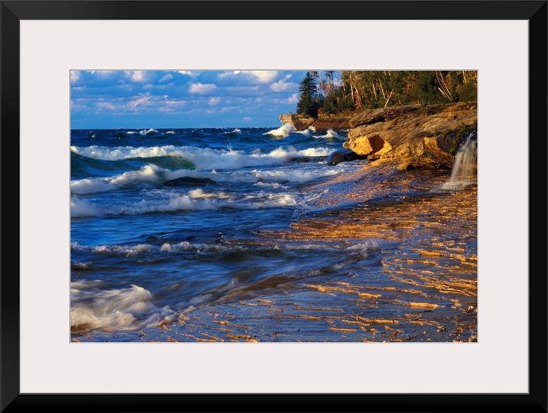 Waves crash onto the rugged lake shore highlighted from the sunset with layered clouds in the distance in this panoramic i...