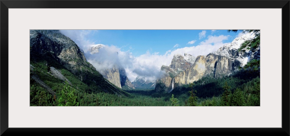 Panoramic photograph shows a valley in Yosemite National Park filled with a dense forest of trees that is surrounded by sn...