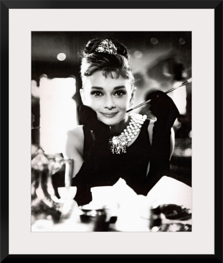 Classic iconic photograph of Audrey Hepburn sitting at a table holding a cigarettte and smiling.