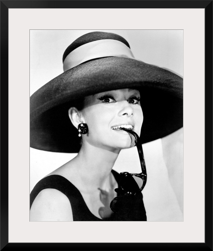 Large photograph of Audrey Hepburn in a giant sun hat holding sunglasses in her mouth as she glances at the camera in a bl...