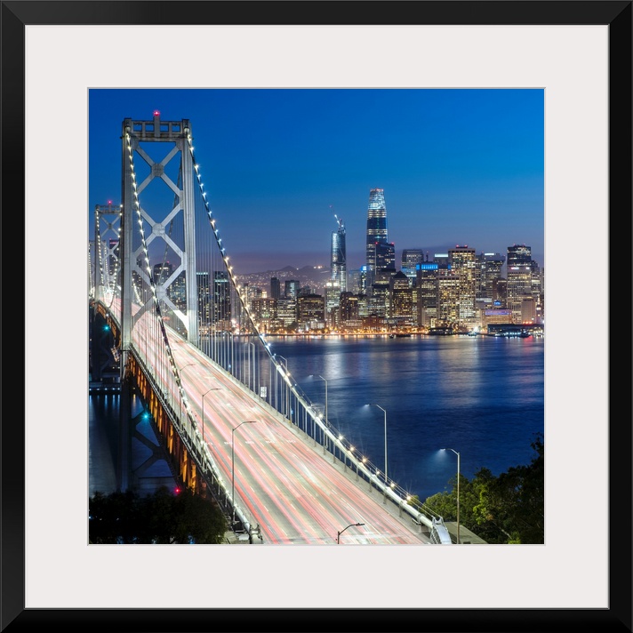 Square photograph of the Bay Bridge at dusk with downtown San Francisco lit up in the background.