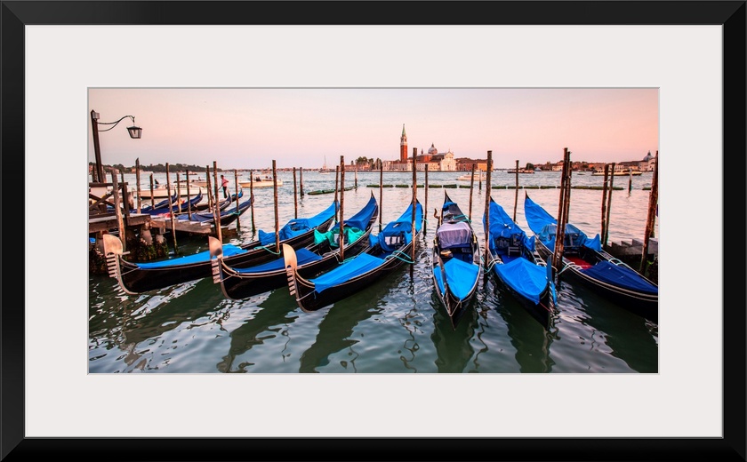 Panoramic photograph of blue gondolas docked in a row on the water with St. Mark's Square in the background at sunset.