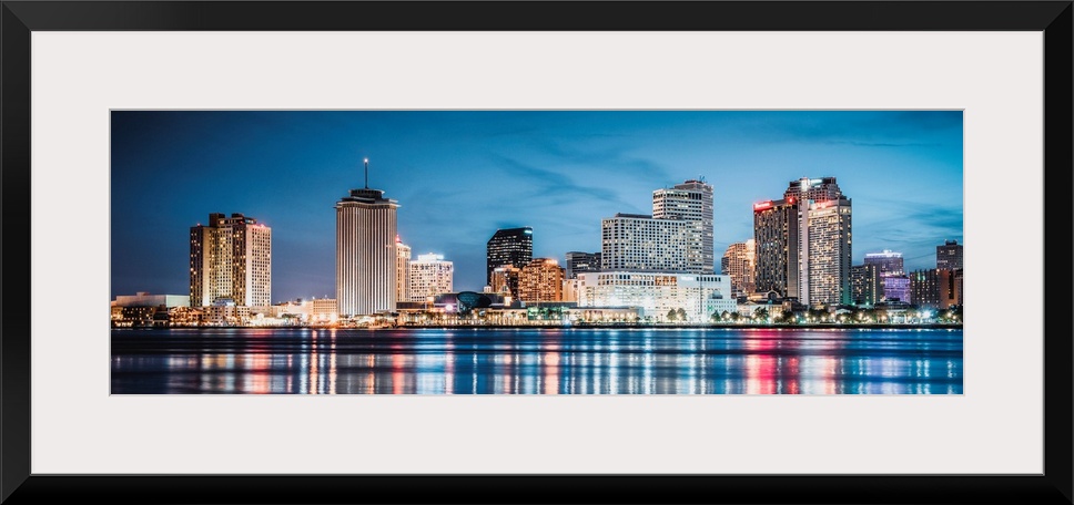 Panoramic photograph of the New Orleans skyline lit up at dusk and reflecting colorful bands onto the Mississippi River.