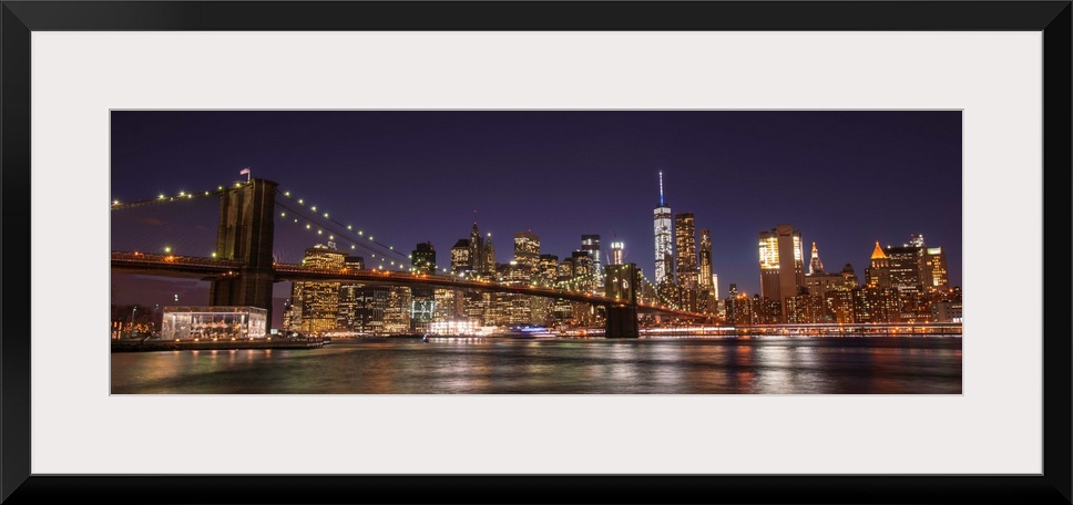 View of the New York City skyline illuminated at night, with the Brooklyn Bridge, from across the water.