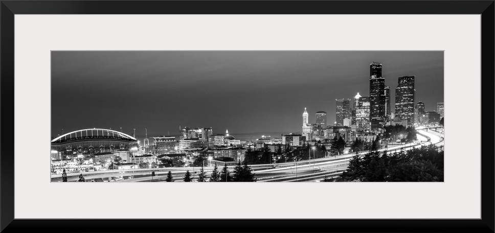 Panoramic photograph of the Seattle skyline with the stadium on the left and light trails from traffic on the highway.
