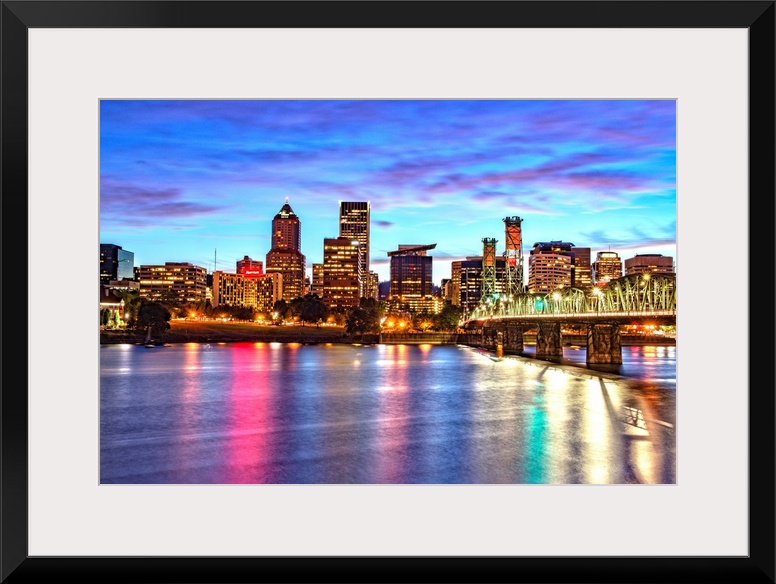 Photograph of the Portland, Oregon skyline lit up at sunset with colors reflecting onto the Willamette River in the foregr...