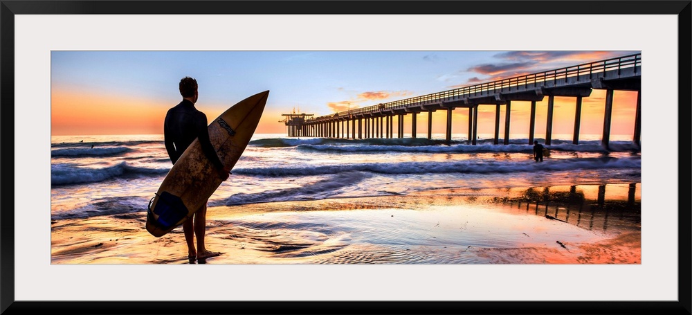 Silhouetted panoramic photograph of a man holding a surf board on the shore of Scripps Beach in San Diego, California, wit...