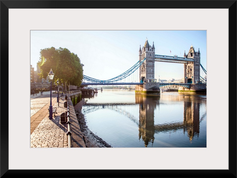 Photograph of Tower Bridge  reflecting on River Thames with a brick sidewalk on the side.