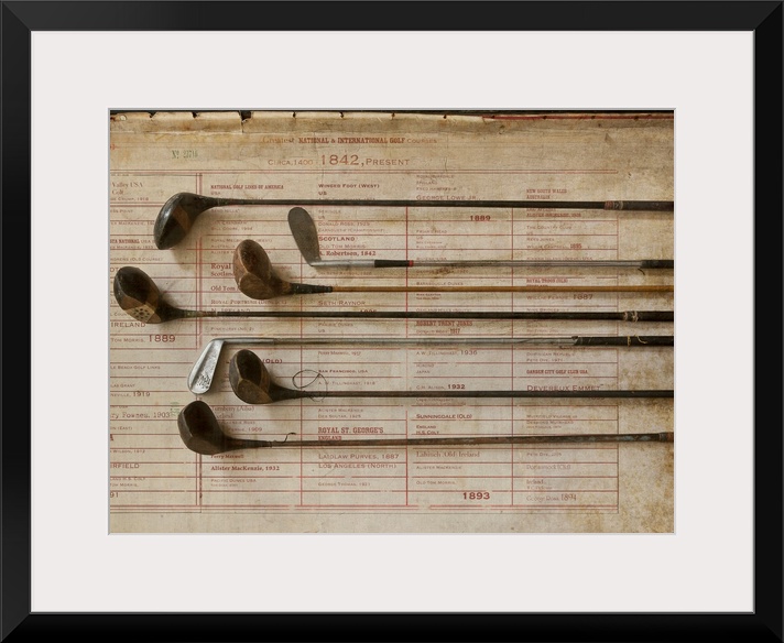 Antique golf clubs are lined up and lay over a vintage poster of major golf tournaments.