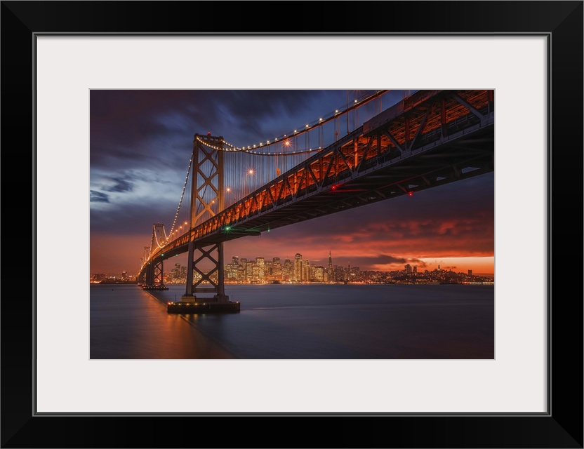 The San Francisco bay bridge illuminated at night with a glowing sunset sky hanging over the city in the distance.