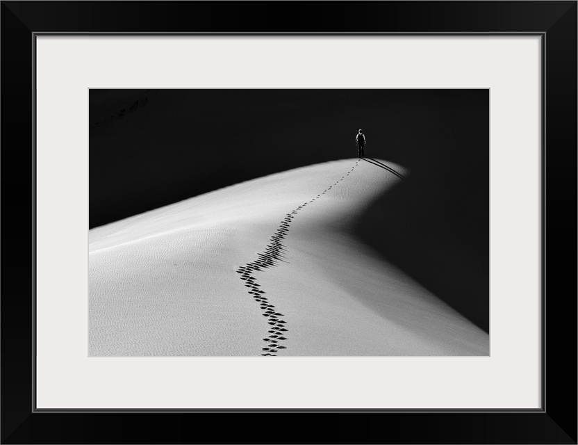 A man walking along the top of a sand dune into darkness leaving footprints behind him.