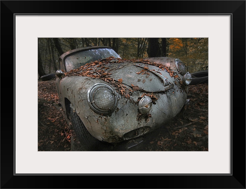 Old, abandoned car in a forest covered in leaves, with cracked, peeling paint.