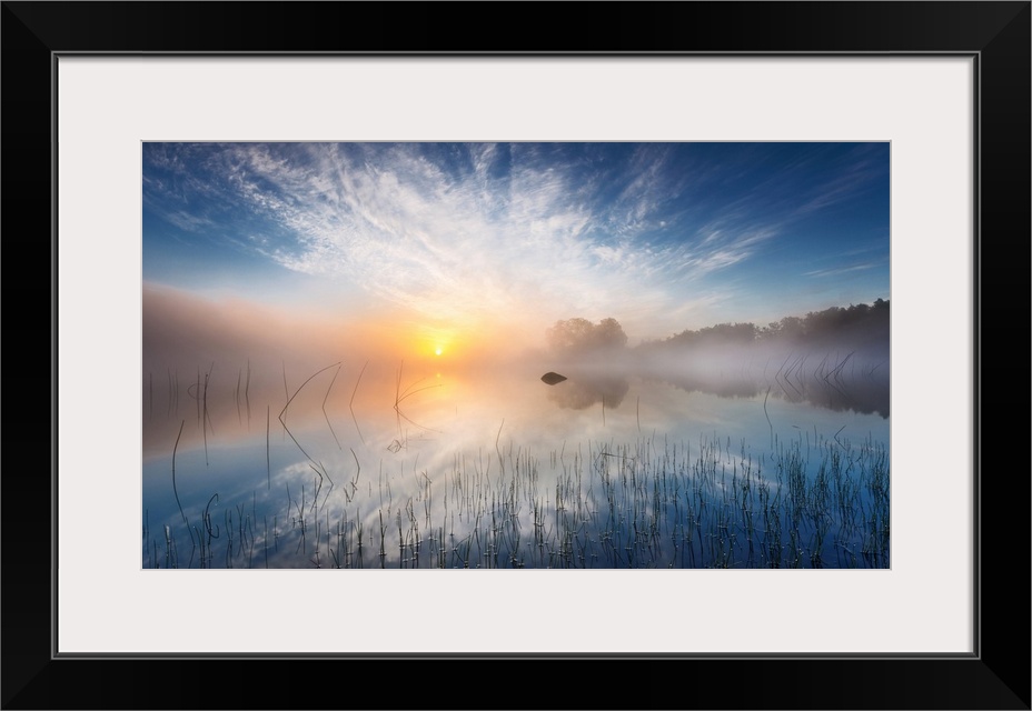 Sunrise in the countryside causing a blanket of fog to appear over the lake the sun and trees are reflected in.