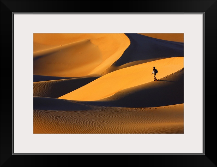 A person walks on a sand dune in a golden desert at sunset.