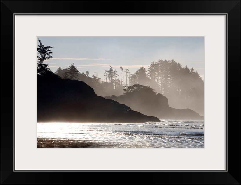Mist rises of the sea against the silhouettes of rocks and trees in this shore line photograph taken in the morning.