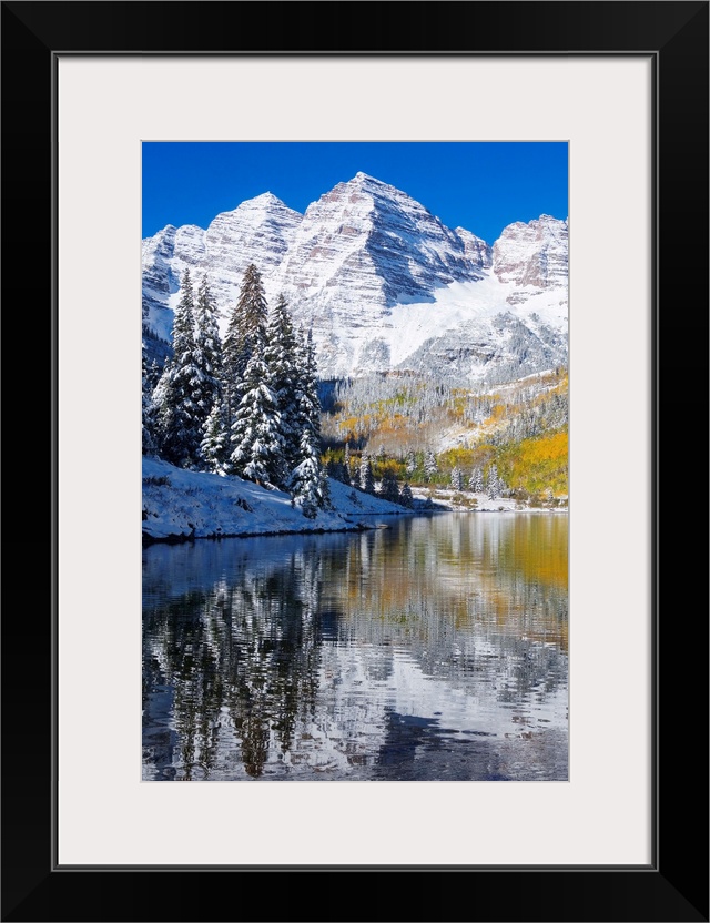 Snow covered mountains are pictured against a clear blue background that reflects down in the body of water that is in the...