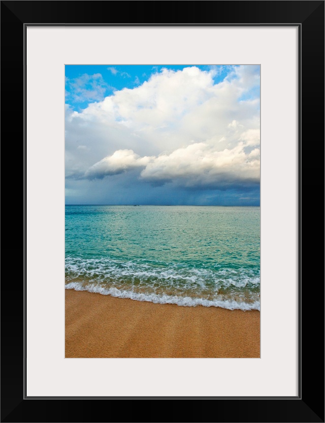 This photograph is taken on a beach in Hawaii of immense clouds that hang in the sky over a teal colored ocean.