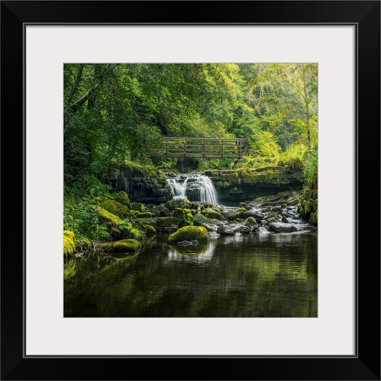 Small waterfall flowing beneath a rustic wooden bridge as it pours over a layered rocky shelf, framed by overhanging trees...