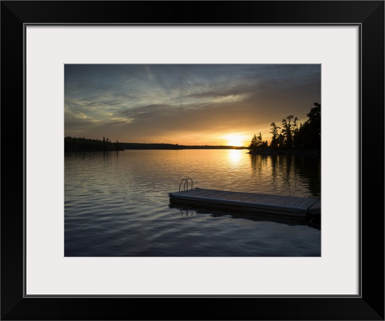 The sun setting over a tranquil lake and silhouetted trees along the shoreline with a dock and ladder in the foreground; L...