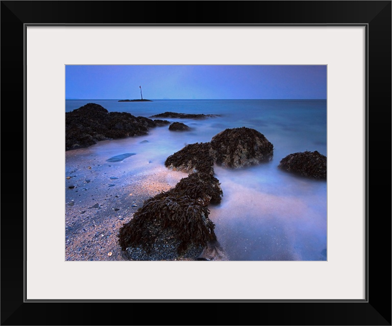 The landscape wall art is a time lapse photograph of waves washing on shore between kelp covered rocks.